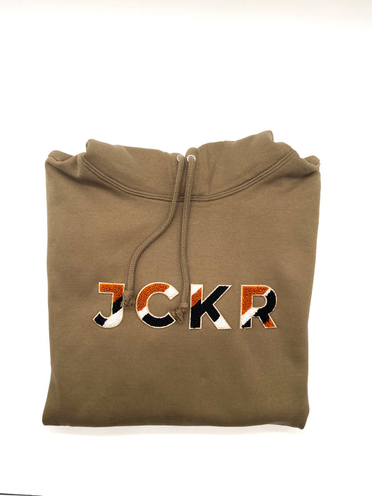 Folded dark beige hoodie with JCKR chenille patch on a white background
