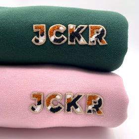Two folded crewneck sweaters, one green and one pink, with JCKR embroidery .