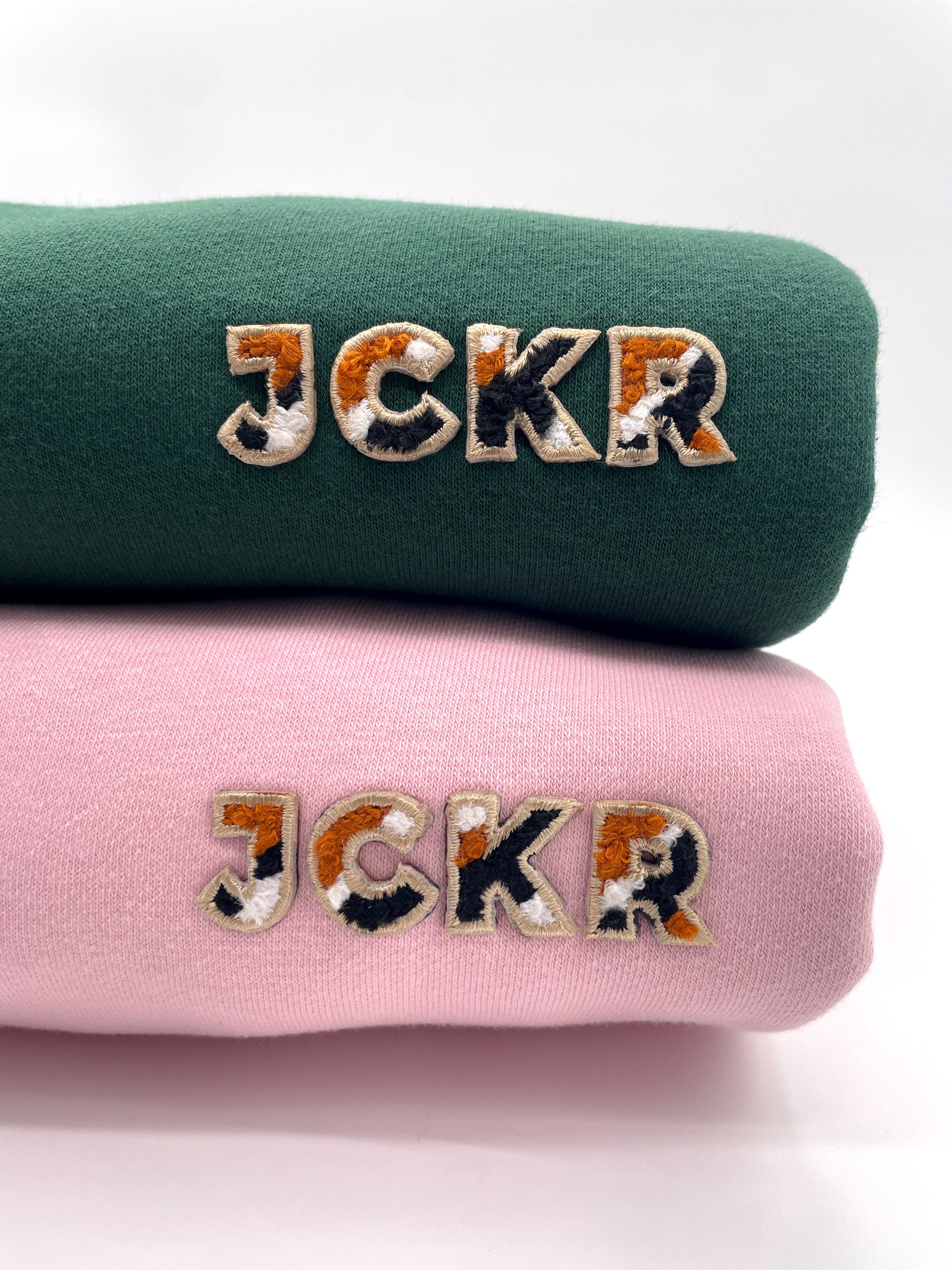 Two folded crewneck sweaters, one green and one pink, with JCKR embroidery .