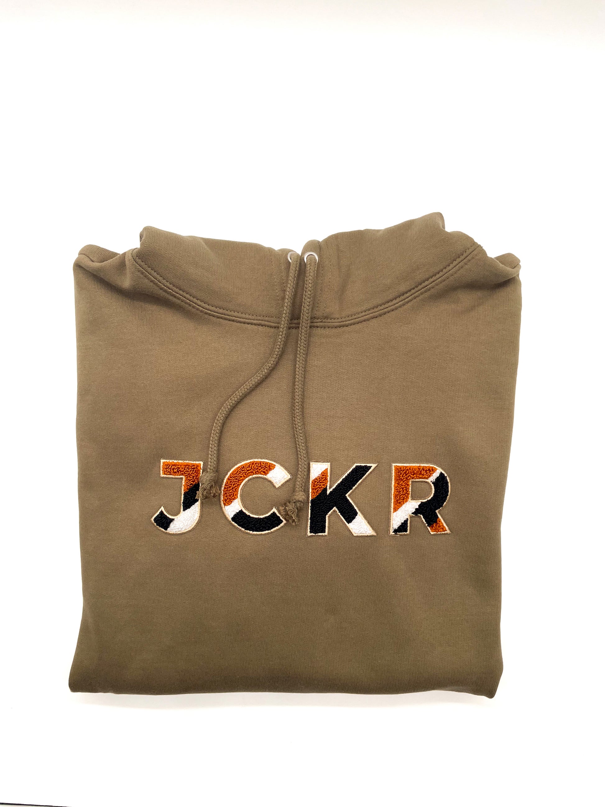 Folded dark beige hoodie with JCKR chenille patch on a white background