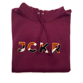 Folded bordeaux hoodie with JCKR chenille patch on a white background