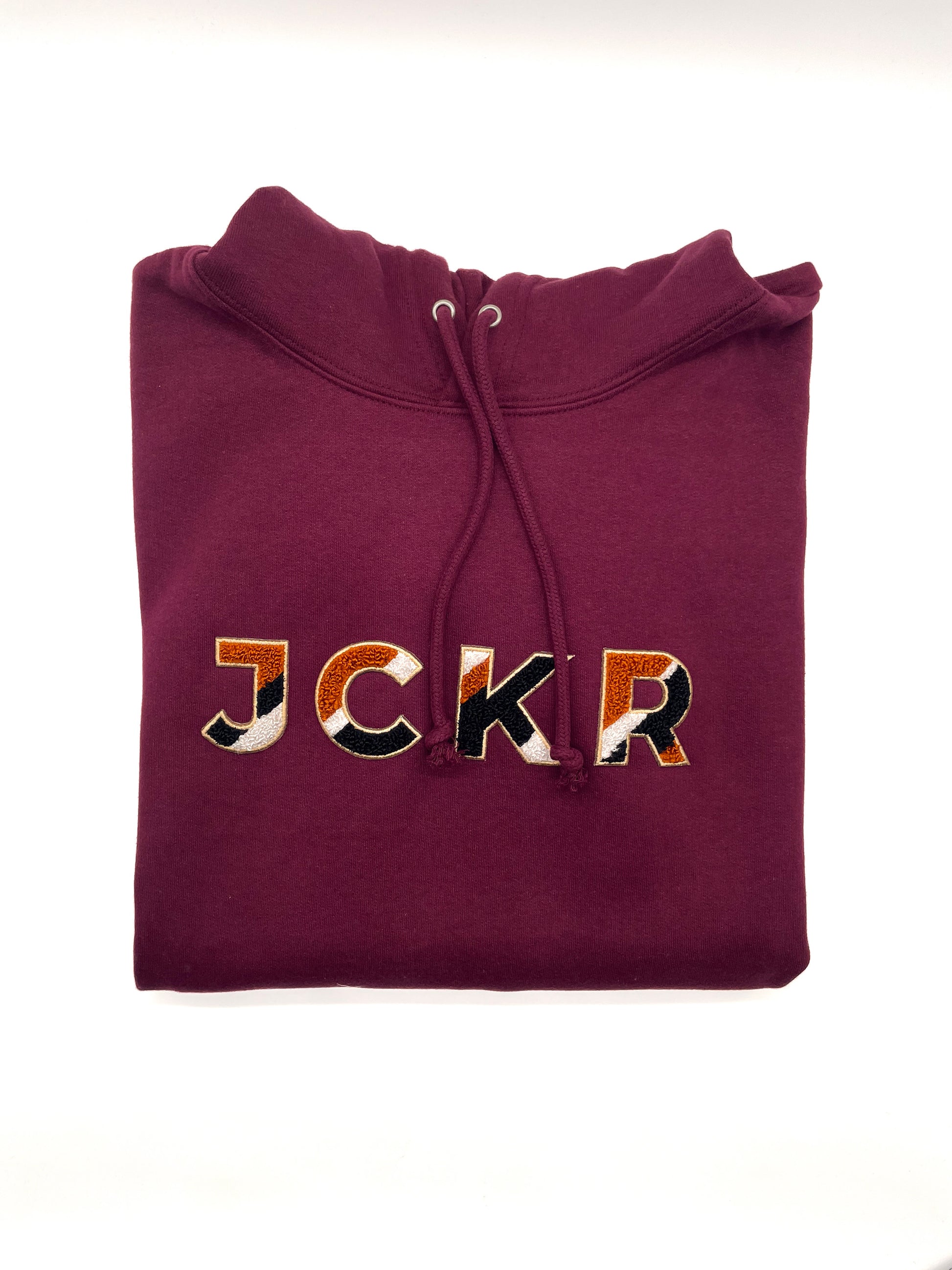 Folded bordeaux hoodie with JCKR chenille patch on a white background