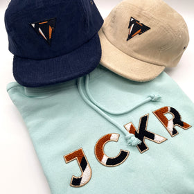 Two 5-panels corduroy hats and a glacial blue hoodie with JCKR chenille patch on a white background