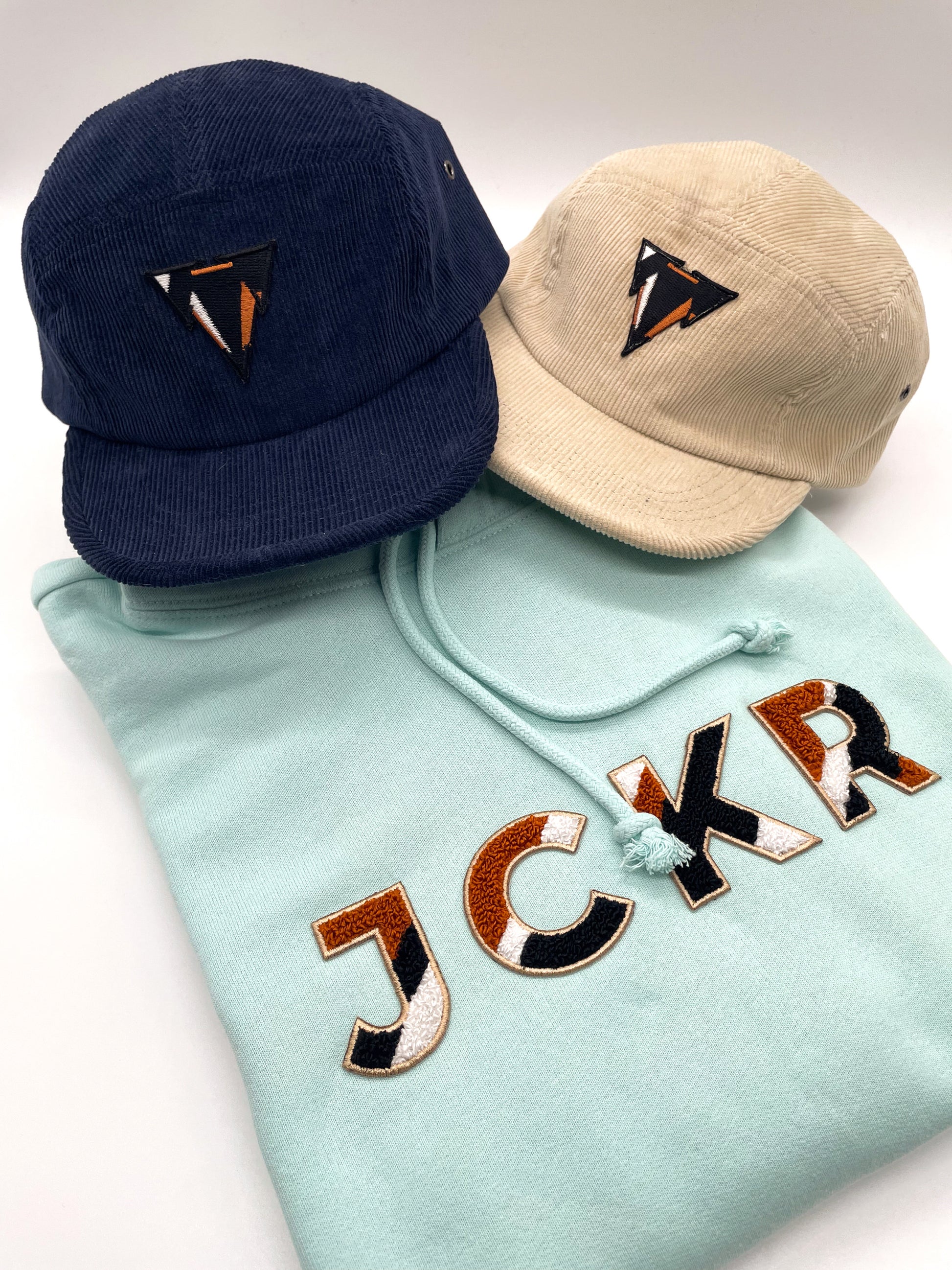 Two 5-panels corduroy hats and a glacial blue hoodie with JCKR chenille patch on a white background