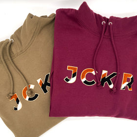 Two hoodies, one bordeaux and one sand beige, with colorful JCKR text design in chenille patch on a white background.