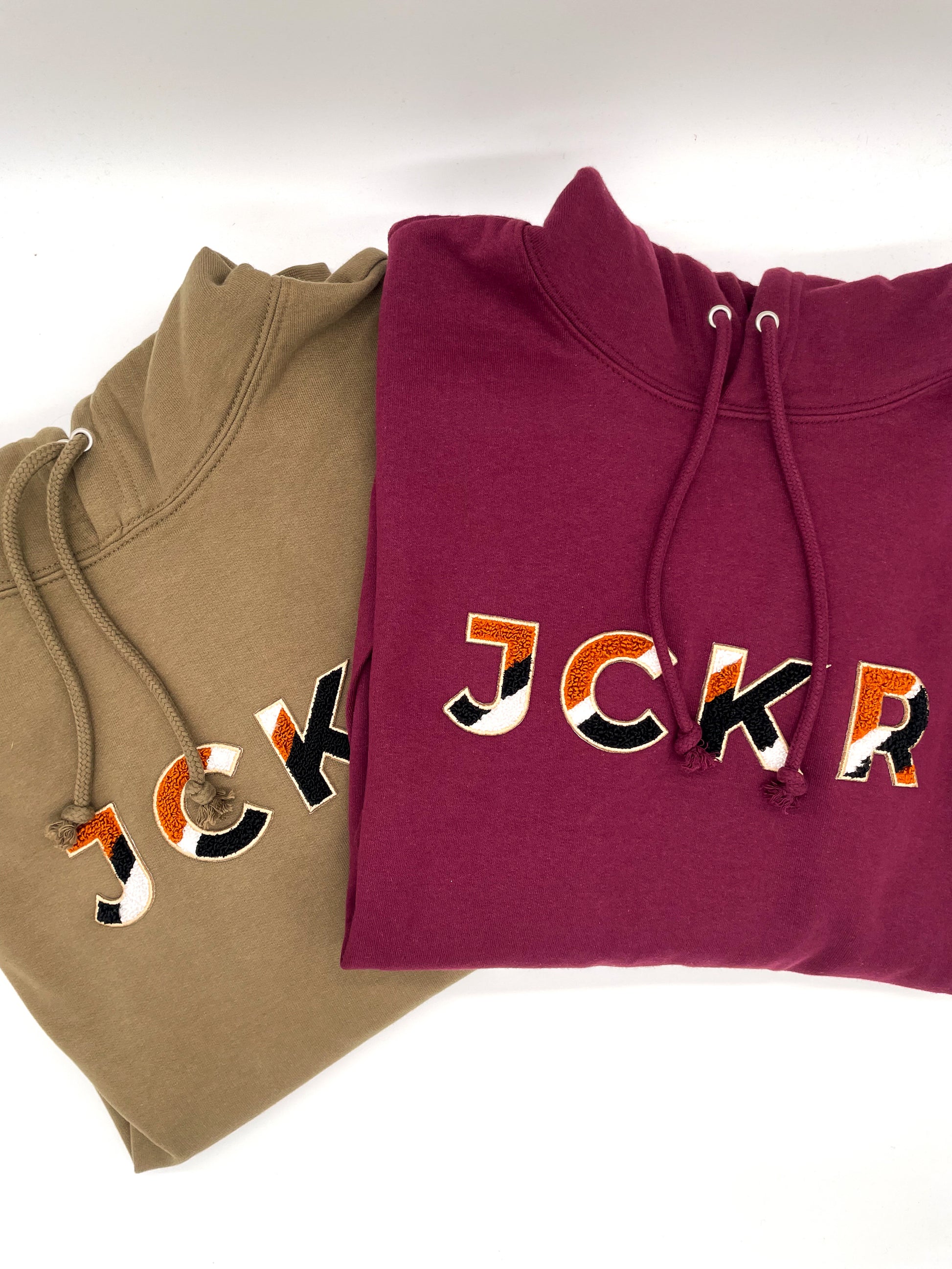 Two hoodies, one bordeaux and one sand beige, with colorful JCKR text design in chenille patch on a white background.