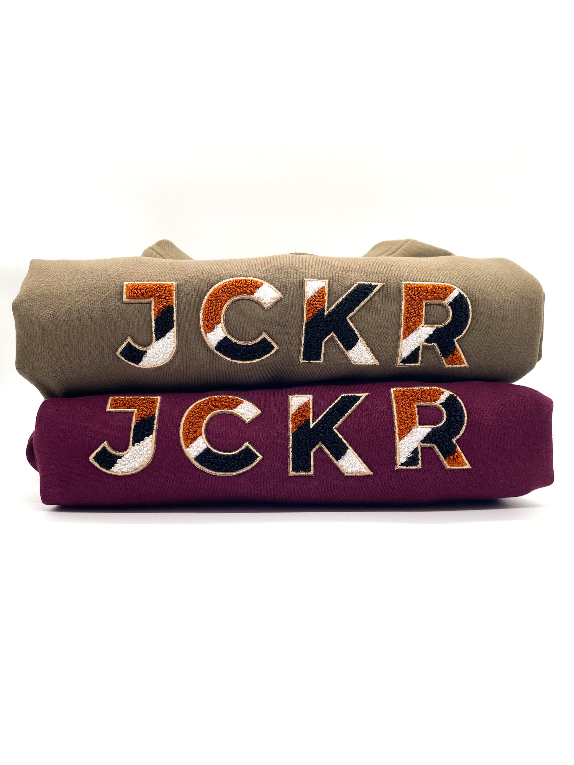 Two folded hoodies with JCKR chenille ptach design on the front, on a white background