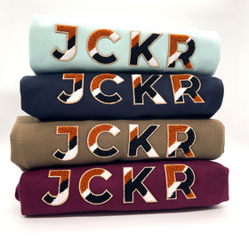 Stack of four folded JCKR winter hoodies with JCKR chenille patch on a white background
