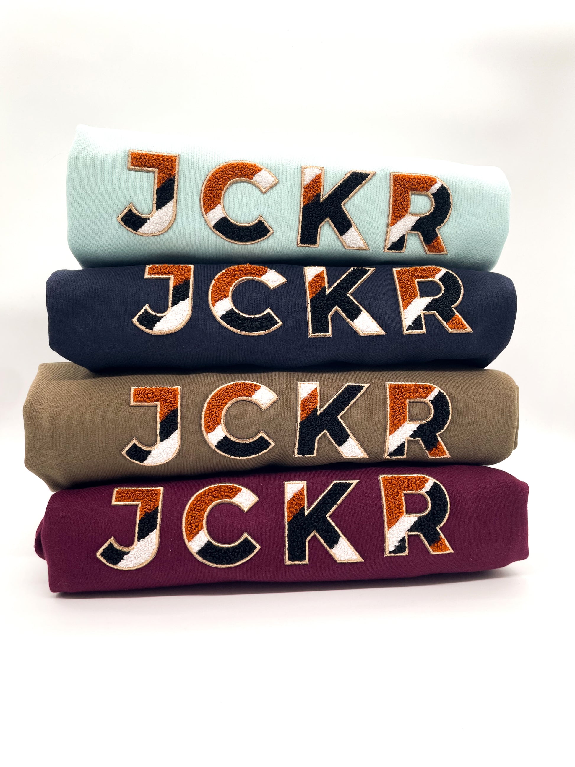 Stack of four folded JCKR winter hoodies with JCKR chenille patch on a white background