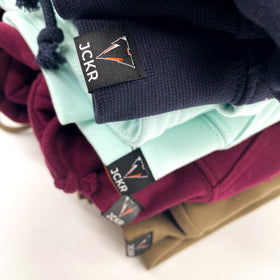 Stack of folded JCKR hoodies with JCKR voewn labels details on a light background