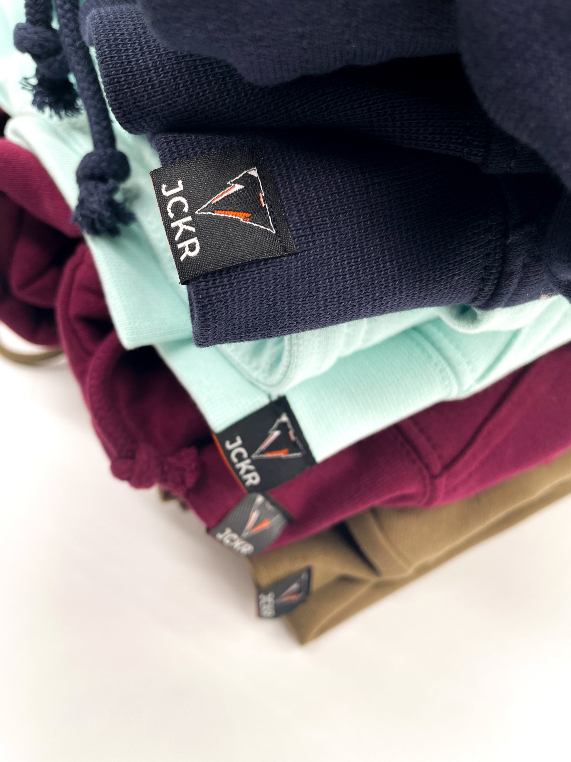 Stack of folded JCKR hoodies with JCKR voewn labels details on a light background
