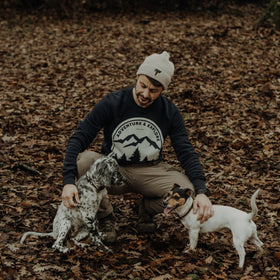 Man sitting on a forest floor with two dogs, wearing a JCKR beanie and 