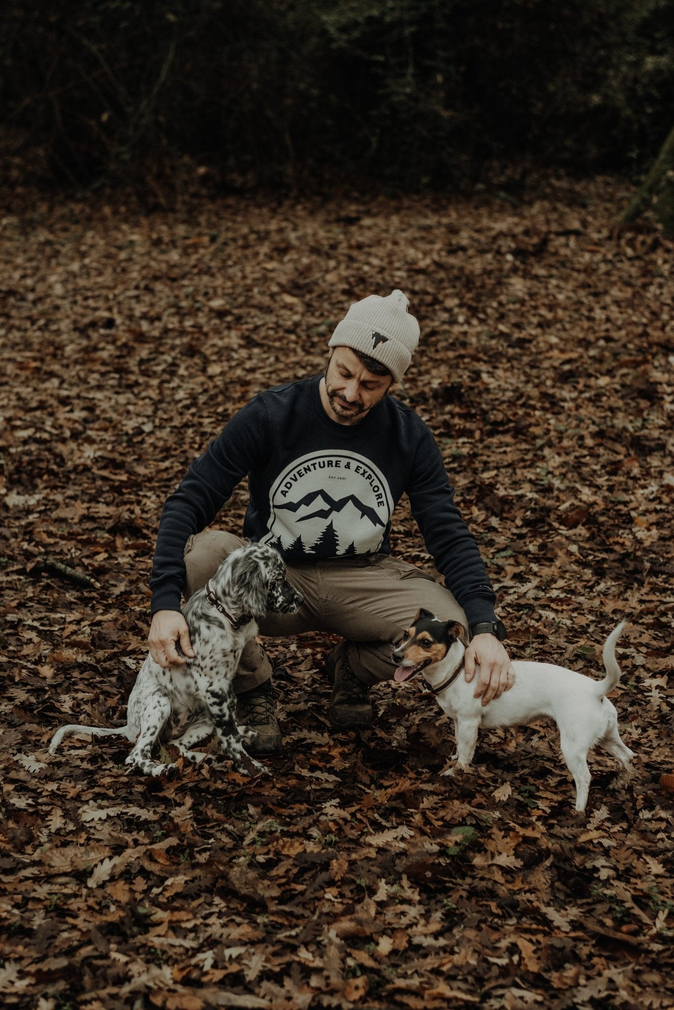 Man sitting on a forest floor with two dogs, wearing a JCKR beanie and "NOVA" sweater.