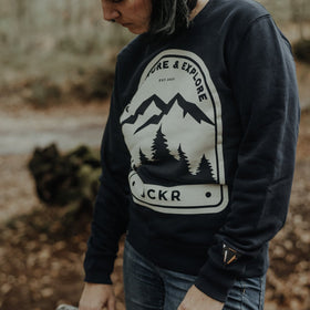 Person wearing a navy NOVA sweatshirt with a mountain and tree design, standing in a forest, from JCKR Clothing