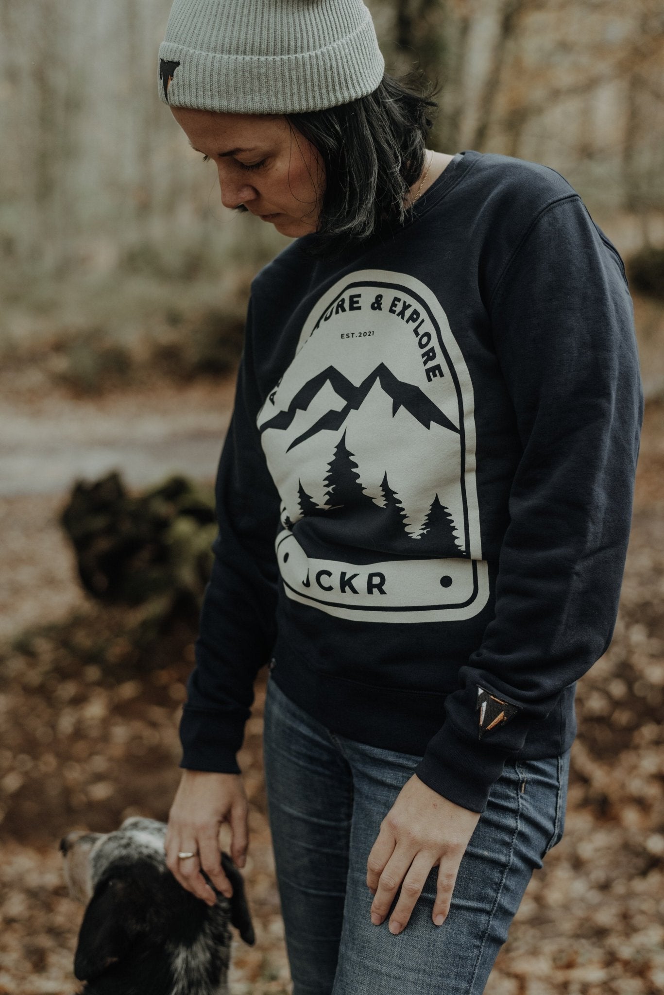 Person wearing a navy NOVA sweatshirt with a mountain and tree design, standing in a forest, from JCKR Clothing