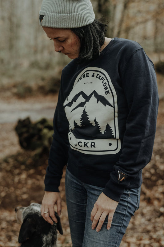 Person wearing a navy NOVA sweatshirt with a mountain and tree design, standing in a forest, from JCKR Clothing