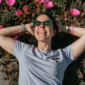 Person wearing sunglasses and a lilac organic cotton SUMMER JCKR t-shirt lying on the ground with pink flowers in the background