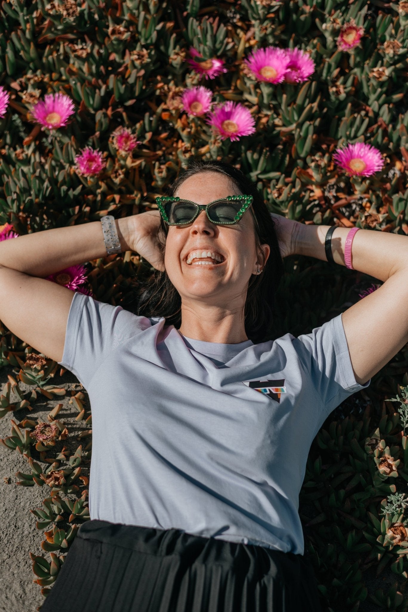 Person wearing sunglasses and a lilac organic cotton SUMMER JCKR t-shirt lying on the ground with pink flowers in the background